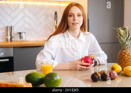 Ritratto di attraente rosso giovane donna seduta al tavolo con frutta tropicale esotica in cucina con interni moderni e luminosi, guardando la macchina fotografica. Foto Stock