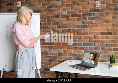 Un'amica lavoratore di mezza età che indossa occhiali in ufficio, guarda lo schermo del laptop, parla con i partecipanti alla conferenza online, speacking a maschio studente, collega Foto Stock