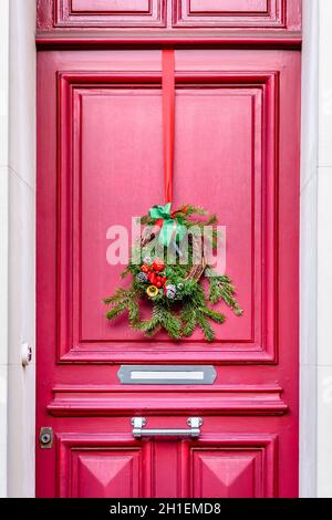 Vista frontale di una corona di Natale fatta di rami di abete, coni di pino e nastri, appesi sulla porta rossa con modanature di una casa cittadina. Foto Stock