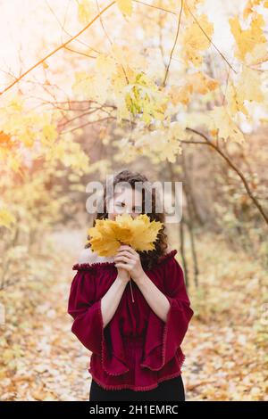 Bella giovane donna con capelli ricci scuri in una parte superiore rossa in un parco autunnale che tiene foglie di acero e sorridente Foto Stock