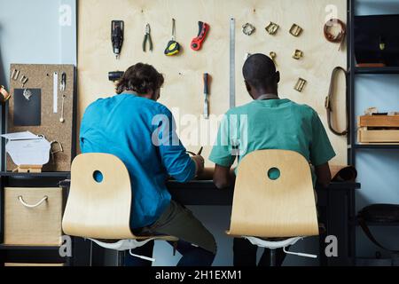 Vista posteriore ritratto di due artigiani maschi che lavorano mentre si siedono da scrivania in laboratorio di pelletteria, spazio copia Foto Stock