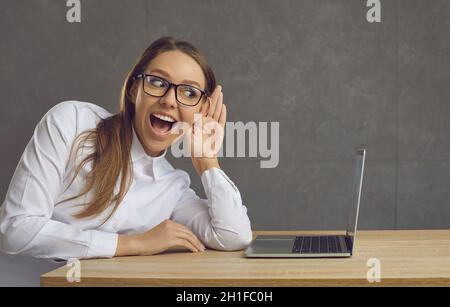 Donna seduta vicino a un portatile cercando di eavesdrop su qualcosa mettendo la sua mano al suo orecchio. Foto Stock