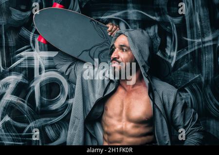 Un latino in una giacca sportiva si erge contro il muro e tiene uno skateboard sulla spalla. Ritratto di un uomo con una longboard. Stile Street. Foto Stock