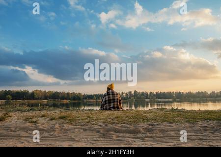 Ragazza viaggiatore in autunno si siede in una coperta plaid sulla riva e guarda il tramonto Foto Stock