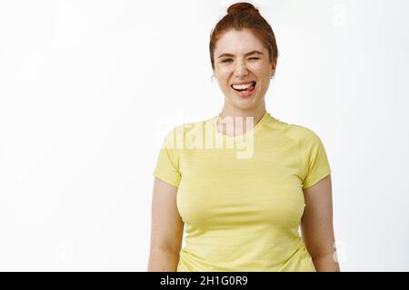 Ritratto di giovane donna entusiasta curvy in vestito di idoneità, mostrando lingua e sorridente, motivato all'allenamento, in piedi su sfondo bianco Foto Stock