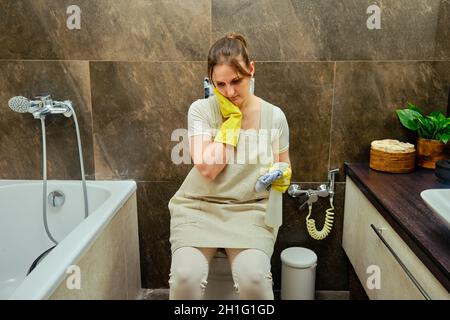 Donna triste si siede sul gabinetto e tiene la testa mentre pulisce il bagno. Disinfezione con guanti di gomma gialli in bagno Foto Stock