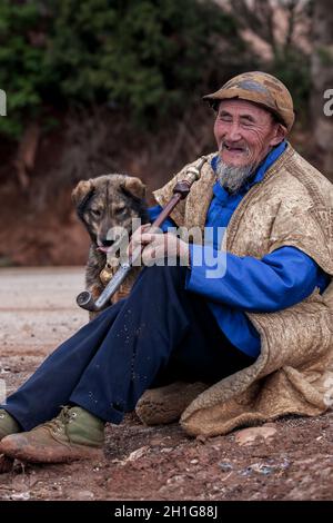 Yunnan, Cina - 5 MARZO 2012: Ritratto di un anziano uomo cinese barbuto con cane sorridente mentre fuma una pipa tradizionale. Yunnan. Foto Stock