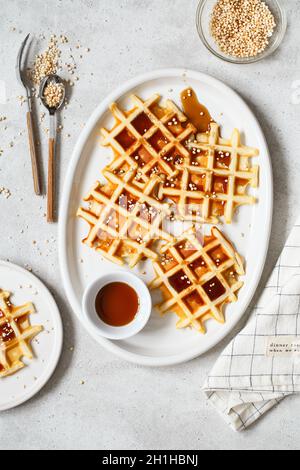 Waffle belgi o di Bruxelles con sciroppo d'acero e quinoa soffiato. Waffle fatti in casa in un piatto ovale bianco sul tavolo da cucina grigio chiaro. Tradizione Foto Stock