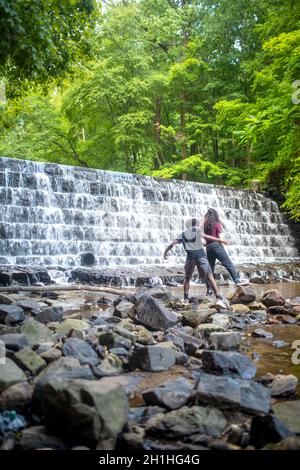 Le cascate d'acqua sopra la cascata a più livelli con lussureggiante vegetazione boschiva alle spalle. Immagine verticale di due giovani, afroamericani e caucasici, alla ba Foto Stock