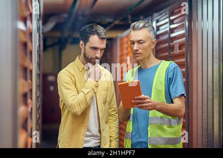 Addetto al magazzino che mostra lo schermo del tablet all'uomo barbato Foto Stock