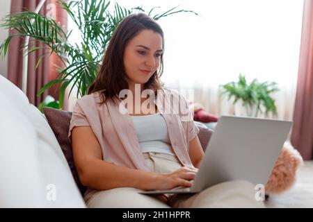 Donna Usa il portatile a casa sul divano. Ritratto giovane 30s calma caucasica donna bruna che lavora in remoto sul portatile o fare shopping online in salotto a. Foto Stock