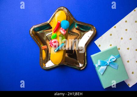 Un piatto a forma di stella d'oro con deliziosa eclair, scatola regalo blu, tovagliolo a forma di cuore bianco giace sul tavolo blu brillante piano. Arredamento festivo per il compleanno, Foto Stock