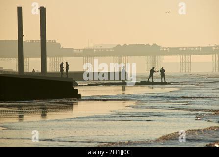 Forme silhoueted di cinque amici su un groyne che conduce al mare a bassa marea. Condivisione selfie. Brighton & Hove, East Sussex, Inghilterra, Regno Unito. Pillars Beach End del vecchio West Pier; Palace Pier dietro. Foto Stock