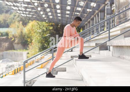 Atleta femminile che fa il fitness in una tuta rosa vicino allo stadio sportivo, African American esegue esercizi di fitness e stretching al mattino Foto Stock