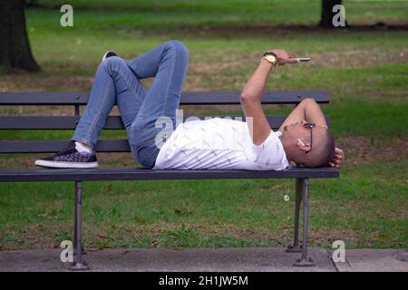 Un uomo di mezza età si trova su una panca che legge il suo telefono cellulare. In un parco a Queens, New York. Foto Stock