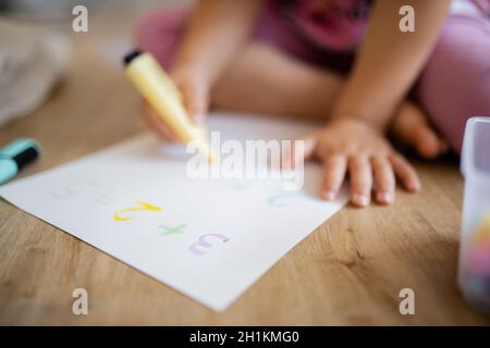 Paesaggio immagine di una bambina in vestiario rosa seduta sul pavimento di legno e fare somme su un foglio di carta con un textmarker giallo, accanto a un Foto Stock