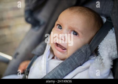 Primo piano di un bambino che piange in abiti grigi e in un passeggino grigio scuro che guarda con tristezza alla fotocamera, con una parete di mattoni sfocati come backgrou Foto Stock