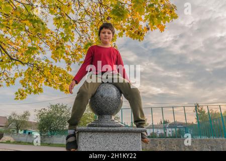 divertente ragazzo in abiti casual si siede su un decorativo recinzione con palla di marmo vicino ad un autunno Foto Stock