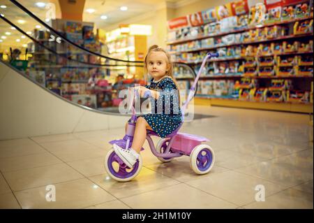 Piccolo e grazioso bambino che ride in bicicletta nel negozio per bambini. Adorabile bambino in attesa di madre in negozio di toyshop, felice infanzia, la ragazza fa un acquisto in capretto Foto Stock