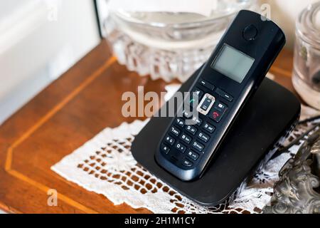 un telefono cordless in plastica nera che si ricarica sulla sua base all'interno di una casa arredata in stile classico con oggetti d'antiquariato e uncinetto bianco. Tecnologia obsoleta Foto Stock