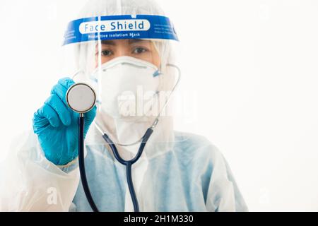 Closeup scienziato medico femminile in uniforme DPI che indossa un viso maschera protettiva e visiera protettiva in plastica che tiene sopra lo stetoscopio medico Mano per controllo Co Foto Stock