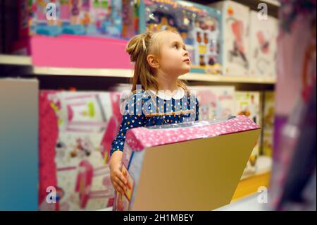 La ragazza piccola graziosa tiene la scatola con la bambola nel deposito del giocattolo. Adorabile bambino che guarda in vetrina in negozio di toyshop, felice infanzia, il bambino fa un acquisto in negozio Foto Stock