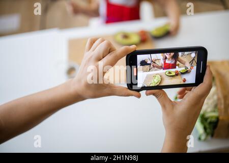 Mani femminili con smartphone registrazione adorabile bambina spargimento avocado su fetta di pane. Carino bambino che taglia le verdure sul tagliere. Foto Stock