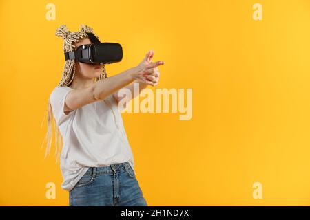 Giovane donna futuristica in t-shirt bianca che tiene le mani come una pistola mentre gioca VR sparatutto su sfondo giallo. Foto Stock