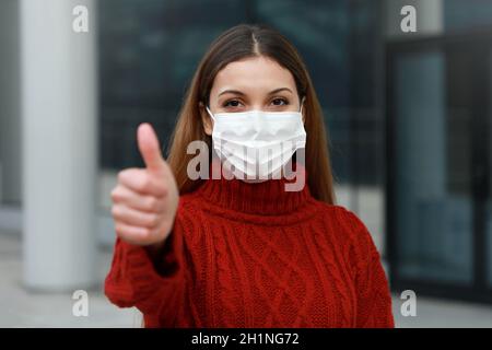 COVID-19 giovane donna ottimista che indossa una maschera protettiva mostrando pollici in su In città strada pronto per la campagna di vaccinazione contro Coronavirus malattie Foto Stock