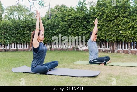 L'uomo e la donna asiatici che praticano lo yoga all'aperto in posizione meditate seduta su erba verde. Giovane coppia che si estende nella natura un campo giardino parco. Med Foto Stock