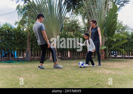 Giovane madre, padre e figlia asiatica giocare a calcio fuori in natura un campo giardino parco. Felice famiglia capretto divertente giocare a calcio insieme in s. Foto Stock