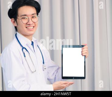 Ritratto closeup di felice asiatico giovane medico bel ragazzo sorridente in uniforme e stetoscopio tracolla che mostra schermo bianco anteriore smart tavolo digitale Foto Stock