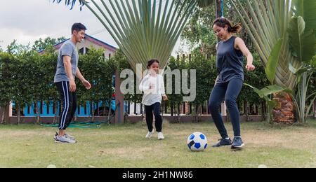 Giovane madre, padre e figlia asiatica giocare a calcio fuori in natura un campo giardino parco. Felice famiglia capretto divertente giocare a calcio insieme in s. Foto Stock