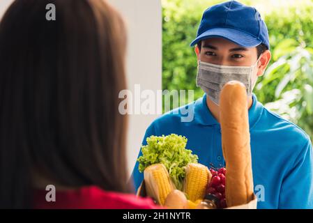 Giovane uomo asiatico di consegna in uniforme indossare maschera protettiva viso che fa servizio di alimentari dando cibo fresco alla donna cliente che riceve front house unde Foto Stock