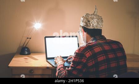 Un giovane uomo con cappuccio in alluminio è seduto nel seminterrato scuro di fronte a un computer portatile. Concetto di teoria della cospirazione Foto Stock