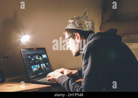 Un giovane uomo con cappuccio in alluminio è seduto nel seminterrato scuro di fronte a un computer portatile. Concetto di teoria della cospirazione Foto Stock