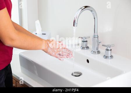 Donna che lavano le mani sotto l'acqua corrente in bagno bianco. Persona che pulisce la pelle con sapone nel lavabo. Femmina con schiuma sulle braccia sotto acqua corrente Foto Stock