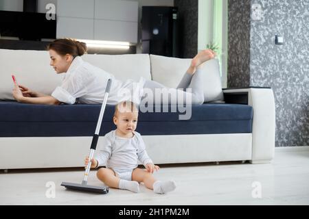 Il bambino gioca con la mop, mentre la madre naviga nel tuo smartphone sdraiato sul divano. Foto Stock