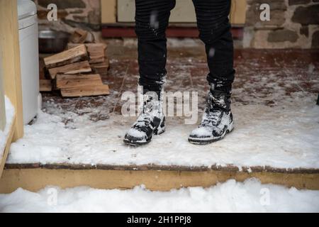 Un giovane in scarponi coperti di neve si erge su un portico innevato da un mucchio di legna da ardere tagliata di fresco. Scena invernale, portico giallo e backgroun casa in pietra Foto Stock