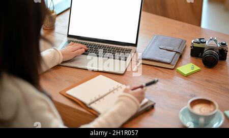 Giovane donna blogger, fotografo freelance che lavora su un computer portatile mentre prende alcune note sul diario personale. Portatile bianco schermo mockup Foto Stock