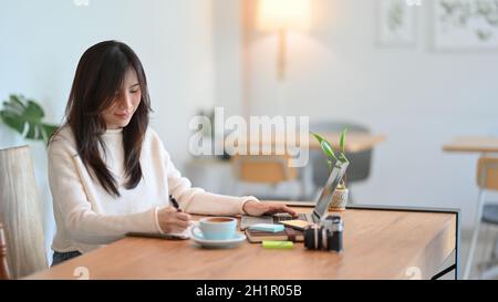 Felice giovane asiatica femmina fare un note sul suo diario mentre si lavora su computer portatile al caffè minimalista Foto Stock