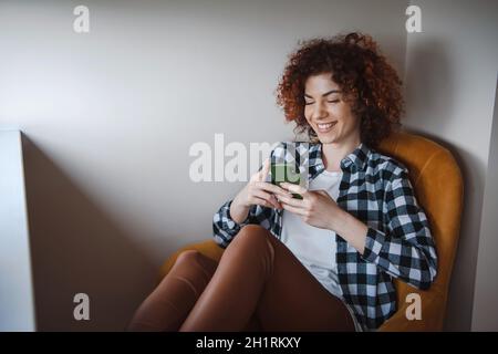 Curly donna caucasica seduta comodamente in poltrona dando messaggi al suo amico che fa il suo sorriso. Faccia sorridente Foto Stock