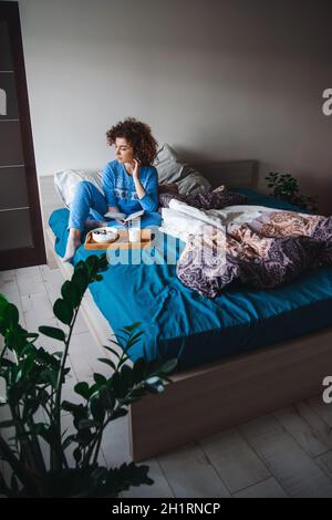 Donna caucasica seduta in pigiama blu a letto guardando indietro e cadendo a pensieri, mentre una sana colazione la attende sul letto. Colazione in camera Foto Stock