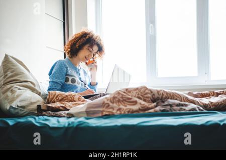 Donna curly che parla al telefono direttamente dal letto notando i compiti. Lezioni online Foto Stock