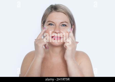 Felice 40s mid invecchiato bionda donna matura applicazione crema facciale sul viso guardando la fotocamera isolato su sfondo bianco. Anti età sana cura della pelle secca Foto Stock