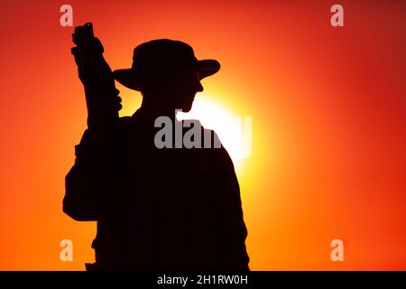 Silhouette di soldati delle forze speciali dell'esercito in boonie e occhiali balistici, in piedi con pistola a corride sullo sfondo del cielo del tramonto. Commando Foto Stock