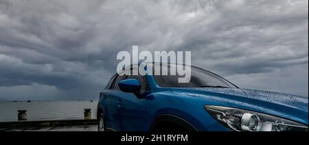 Auto SUV blu con gocce d'acqua parcheggiata nel parcheggio vicino alla spiaggia di mare contro il cielo tempestoso e nuvoloso. Vista frontale della nuova auto SUV di lusso con design sportivo. Foto Stock