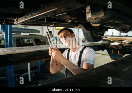 Ritratto medio di serio bel meccanico professionale maschile auto in uniforme in piedi in buca di ispezione e di lavoro con l'utensile. Foto Stock