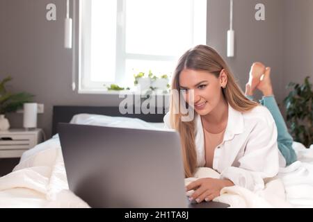Bella ragazza caucasica sorridente si trova sul letto in camera da letto e utilizzare il computer portatile per il lavoro remoto. Concetti di comunicazione, tecnologia, lavoro a distanza in interni grigi Foto Stock