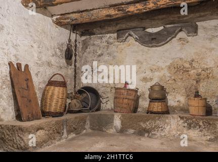 Seminterrato di una vecchia casa con travi in legno e muro con umidità, oggetti antichi della casa, carboy demijohn, pentola, asse, Cestello vimini, puleggia Foto Stock